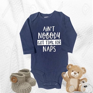 May include: A navy blue long-sleeved baby onesie with white text that reads "Ain't Nobody Got Time For Naps". The onesie is paired with a pair of grey knitted baby booties and a brown teddy bear.