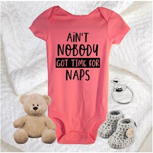 May include: A pink baby onesie with the text "Ain't Nobody Got Time For Naps". The onesie is next to a teddy bear, a silver baby ring, and a pair of grey knitted baby booties.