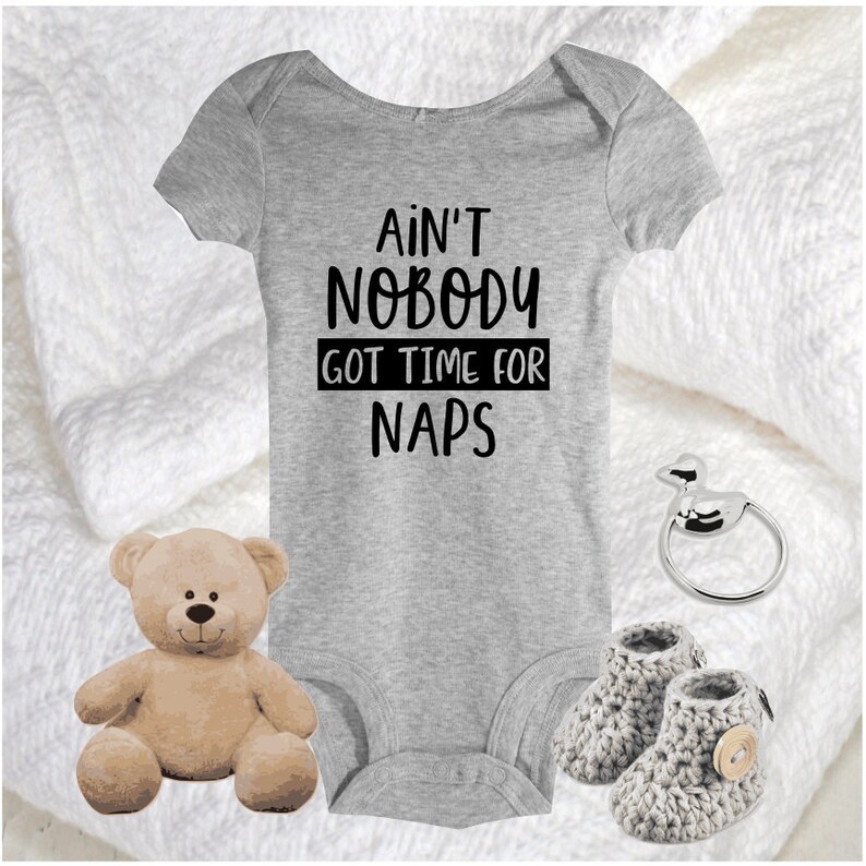 May include: A grey baby onesie with the text "Ain't Nobody Got Time For Naps" in black. The onesie is on a white surface with a teddy bear, a pair of grey booties, and a silver baby rattle.