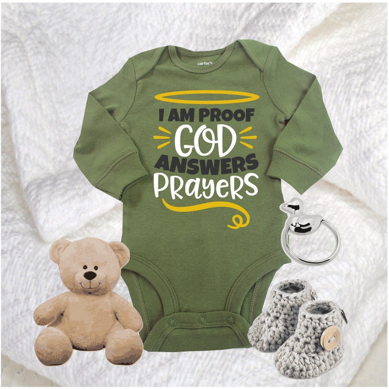 May include: A green long-sleeved baby onesie with the text "I am proof God answers prayers" in yellow and black lettering. The onesie is on a white fuzzy blanket with a teddy bear and a silver baby rattle.