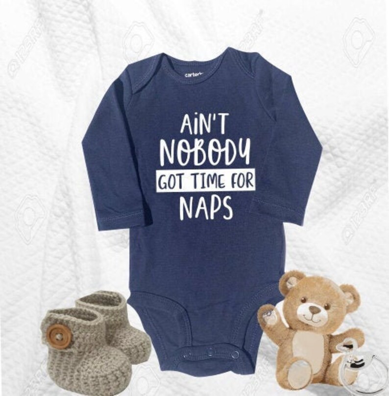 May include: A navy blue long-sleeved baby onesie with white text that reads "Ain't Nobody Got Time For Naps". The onesie is paired with a pair of grey knitted baby booties and a brown teddy bear.