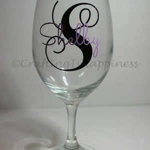 May include: A clear glass wine glass with a black and purple vinyl decal that says "Shelby" with a decorative swirl.