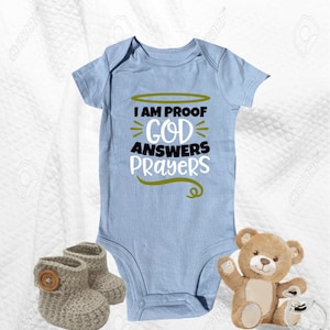 May include: A light blue baby onesie with the text "I am proof God answers prayers" in black and gold lettering. The onesie is on a white surface with a brown teddy bear and two gray knitted booties.