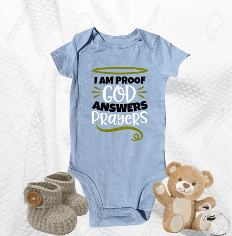 May include: A light blue baby onesie with the text "I am proof God answers prayers" in black and gold lettering. The onesie is on a white surface with a brown teddy bear and two gray knitted booties.