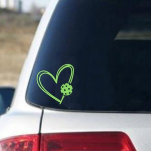May include: A green heart-shaped sticker with a green and white patterned circle inside. The sticker is on a car window.