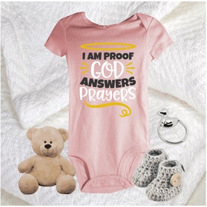 May include: A pink baby onesie with the text "I am proof God answers prayers" in black and gold lettering. The onesie is surrounded by a white blanket, a teddy bear, a silver baby rattle, and a pair of gray knitted baby booties.