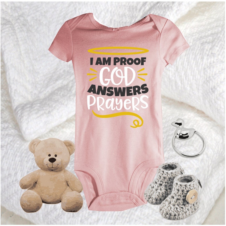 May include: A pink baby onesie with the text "I am proof God answers prayers" in black and gold lettering. The onesie is surrounded by a white blanket, a teddy bear, a silver baby rattle, and a pair of gray knitted baby booties.