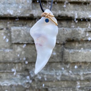 May include: A white, irregularly shaped pendant necklace. The pendant is adorned with a gold-colored wire wrap and a small blue gemstone. The necklace hangs from a black cord, creating a simple yet elegant design.