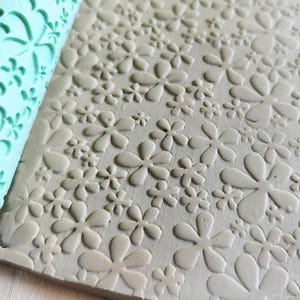 May include: A mint green silicone pottery texture roller featuring a repeating pattern of various sized five-petaled flowers. The roller is shown pressed into a flat slab of grey clay, leaving a clear, embossed floral impression across the surface.