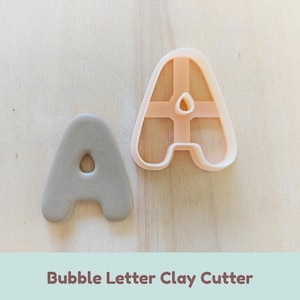 Puede incluir: Un cortador de arcilla de plástico color melocotón claro con forma de letra A, con una estructura de soporte transversal y un recorte de arcilla gris a juego, que presenta un diseño de fuente redondeada con una pequeña abertura central en forma de gota.