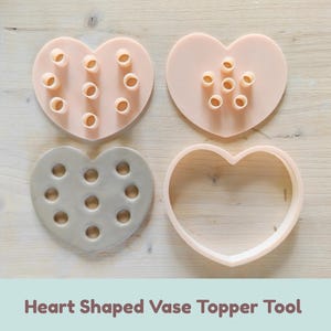 Pode incluir: Um conjunto de ferramentas em forma de coração para o topo de vasos, incluindo duas placas de plástico em tom pêssego claro com tubos circulares salientes, uma placa cinzenta com recortes circulares e uma moldura em forma de coração, rotulado Heart Shaped Vase Topper Tool.
