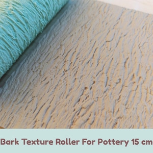May include: A light blue cylindrical pottery texture roller with a bark pattern, positioned next to a flat, grey clay slab that displays the same embossed tree bark texture. The tool is labeled as a Bark Texture Roller For Pottery 15 cm.