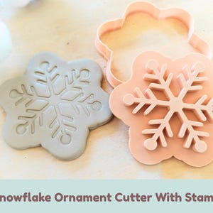 Pode incluir: Um cortador de biscoitos em forma de floco de neve na cor pêssego claro e um carimbo de floco de neve cinza correspondente. O carimbo apresenta um design detalhado de floco de neve de seis pontas com pontas arredondadas. O texto "Snowflake Ornament Cutter With Stamp" está impresso abaixo.