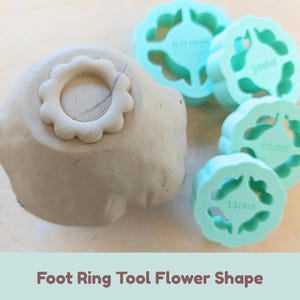 Può includere: Un set di quattro strumenti per ceramica color verde menta a forma di fiore per anelli di base, con misure 35mm, 40mm, 50mm e 60mm in rilievo. Uno strumento è premuto in una forma di argilla grigia, creando un'impronta. Testo: Foot Ring Tool Flower Shape.