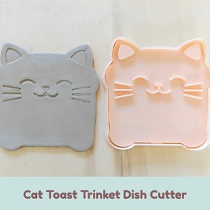 Può includere: Un timbro grigio a forma di gatto e un tagliabiscotti rosa chiaro a forma di gatto. Entrambi gli oggetti presentano una testa quadrata con orecchie a punta, occhi chiusi, un naso piccolo e tre baffi per lato. Il testo recita Cat Toast Trinket Dish Cutter.