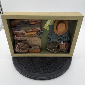 Puede incluir: Una caja decorativa con temática de pesca. Incluye un pez, caña de pescar, chaleco, sombrero, red y otros accesorios. La caja tiene un marco verde claro y una base negra.