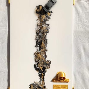 Puede incluir: Obra de arte rectangular blanca con detalles dorados en las esquinas. Un pequeño coche gris está situado en la parte superior, sobre un diseño fluido de resina negra, dorada y blanca. Un emblema dorado y texto en la parte inferior.