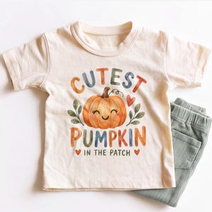 May include: A cream-colored short-sleeve t-shirt featuring a watercolor-style graphic of a smiling orange pumpkin with green leaves and the text "CUTEST PUMPKIN IN THE PATCH" in multi-colored letters, paired with sage green ribbed pants.