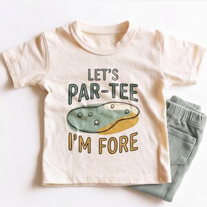 May include: A cream-colored short-sleeve t-shirt featuring the text LET'S PAR-TEE I'M FORE in dark green and yellow, with a graphic of a golf green and golf balls. Beside the shirt are sage green corduroy pants with an elastic waistband.