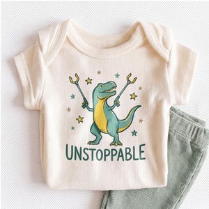 May include: A cream-colored ribbed short-sleeve bodysuit featuring a graphic of a green and yellow dinosaur holding two grabber tools, surrounded by small stars, with the text UNSTOPPABLE printed in dark green capital letters below the dinosaur.