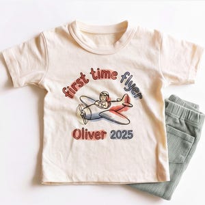 May include: A cream-colored short-sleeve t-shirt featuring a graphic of a pilot in a small airplane with the text "first time flyer" and "Oliver 2025" in red and blue. The shirt is paired with sage green ribbed pants with an elastic waistband.