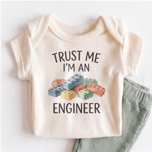 May include: A cream-colored short-sleeve bodysuit featuring the black text TRUST ME I'M AN ENGINEER above a graphic of colorful plastic building blocks in shades of blue, green, orange, and yellow. The garment has a ribbed texture and short sleeves.