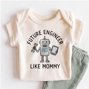 May include: A cream-colored short-sleeve baby bodysuit featuring a graphic of a grey robot holding a wrench and a tablet. The text "FUTURE ENGINEER LIKE MOMMY" is printed in black above and below the robot illustration. The fabric has a ribbed texture.
