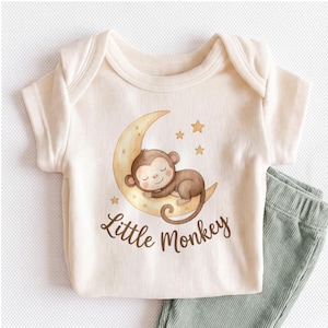 May include: A cream-colored ribbed short-sleeve bodysuit featuring a graphic of a brown monkey sleeping on a yellow crescent moon surrounded by small stars. The text Little Monkey is printed in brown script below the moon illustration.