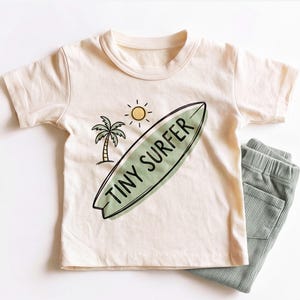 May include: A cream-colored short-sleeve t-shirt featuring a graphic of a green surfboard with the text TINY SURFER, a palm tree, and a yellow sun. Beside the shirt are sage green ribbed pants with an elastic waistband and side pockets.