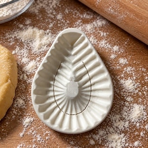 May include: A white, teardrop-shaped cookie mold featuring a central raised oval surrounded by radiating, fluted ridges. The mold has a smooth, matte finish and a distinct, symmetrical pattern designed to imprint dough with a decorative texture.