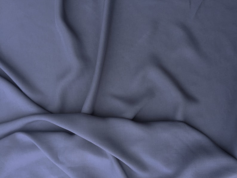 May include: A close-up of a draped fabric in a muted blue-purple shade. The material features soft folds and subtle shadows, creating depth and texture. The fabric appears smooth and flowing, suggesting a lightweight material.