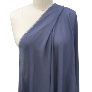 May include: A length of blue fabric with a frayed edge, draped over a mannequin. The fabric is a solid, muted blue colour and appears lightweight. The material is arranged to show its drape and texture. The fabric's edge has a raw, fringed finish.