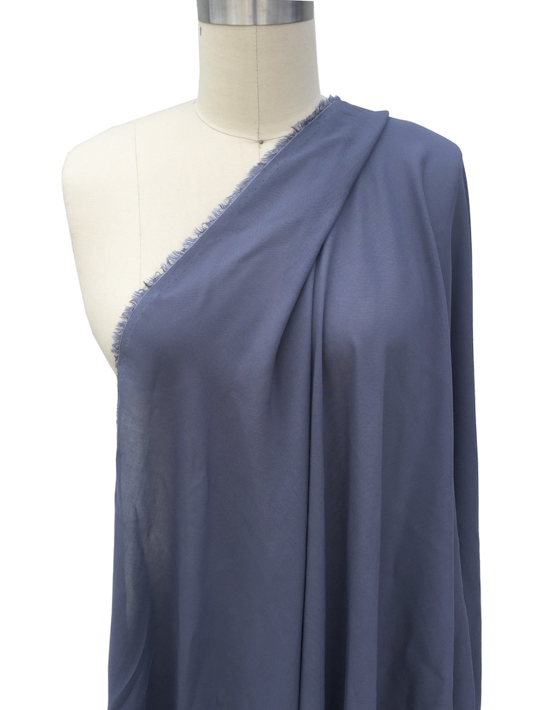 May include: A length of blue fabric with a frayed edge, draped over a mannequin. The fabric is a solid, muted blue colour and appears lightweight. The material is arranged to show its drape and texture. The fabric's edge has a raw, fringed finish.