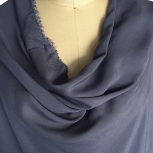 May include: A draped, slate blue fabric is displayed on a mannequin. The fabric has a soft texture and a cowl neckline. The edge of the fabric has a frayed, unfinished detail. The fabric is a solid colour.