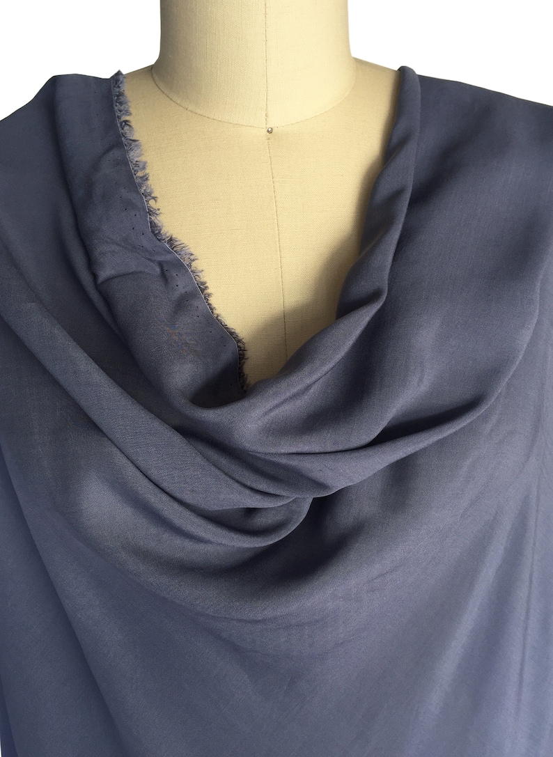 May include: A draped, slate blue fabric is displayed on a mannequin. The fabric has a soft texture and a cowl neckline. The edge of the fabric has a frayed, unfinished detail. The fabric is a solid colour.