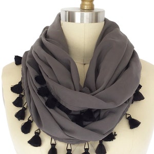 May include: A grey infinity scarf with black tassels. The scarf is draped around a mannequin, showing its soft, flowing fabric and the decorative tassels along the edges. This accessory is a stylish addition to any wardrobe.