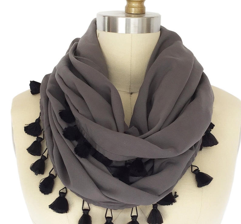 May include: A grey infinity scarf with black tassels. The scarf is draped around a mannequin, showing its soft, flowing fabric and the decorative tassels along the edges. This accessory is a stylish addition to any wardrobe.