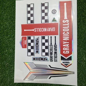 Può includere: Un foglio di adesivi per mazze da cricket con il marchio "GRAY-NICOLLS" in rosso e bianco. Gli adesivi presentano motivi a scacchi, disegni a freccia e la parola "DELTA". Il foglio è su una superficie verde.