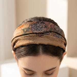 May include: A brown floral headband with a vintage aesthetic. The headband features a repeating pattern of blue, purple, and orange flowers with small gold-colored embellishments. The fabric appears to be soft and slightly textured.