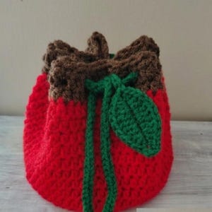 Peut inclure: Un sac à cordon fait à la main en crochet en forme de fraise. Le sac est principalement rouge avec un dessus marron et une feuille et un cordon de serrage verts. Le sac est fait de fil et a un aspect texturé.