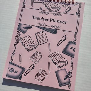 May include: A pink Teacher Planner with a copper spiral binding. The cover features the text "Teacher Planner" in a white banner, surrounded by black illustrations of books, pens, and other school-related items. The planner is designed for teachers.