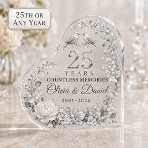 May include: A heart-shaped clear acrylic plaque featuring the text 25 YEARS COUNTLESS MEMORIES, Olivia & Daniel, and 2001-2026. The design includes two birds, floral arrangements, and pearl-like accents in silver and white tones.
