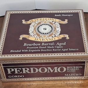 May include: A rectangular Perdomo Habano cigar box with a dark brown finish. The box features gold accents and text that reads "Bourbon Barrel-Aged" and "Premium Hand-Made Cigars." The box is labeled "Gordo Maduro" and "Esteli, Nicaragua."
