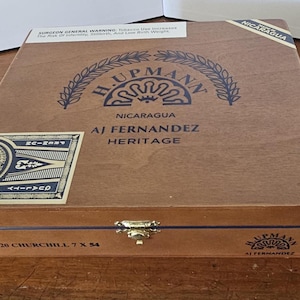 May include: A wooden cigar box with the text "H. Upmann Nicaragua AJ Fernandez Heritage" in dark blue. The box has a gold clasp and a label with the text "1844" and "Quality". The box is labeled "20 Churchill 7 x 54".