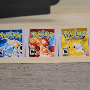 May include: Three Pokemon Game Boy cartridges. The cartridges feature the Pokemon logo and character art. The first cartridge is blue with Blastoise, the second is red with Charizard, and the third is yellow with Pikachu. The ESRB rating is visible.