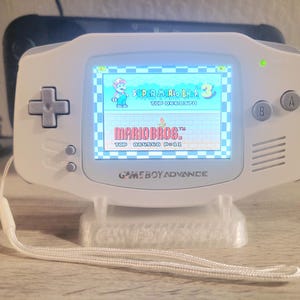 Puede incluir: Una consola portátil Game Boy Advance blanca que muestra el juego Super Mario Bros. 3. La pantalla muestra un nivel colorido con Mario. La consola está sobre un soporte blanco con un cordón blanco. El texto "MARIO BROS" es visible.