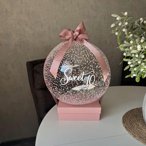 Op de afbeelding: Een heldere, ronde ballon gevuld met roze confetti en de tekst "Sweet 10". De ballon is vastgebonden met een roze lint en staat op een roze doos. Een decoratief cadeau voor een tiende verjaardag.