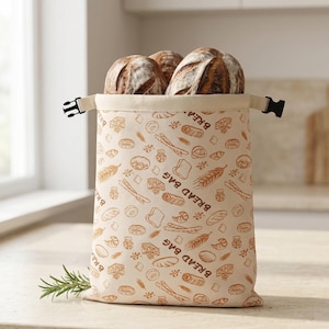Könnte beinhalten: Beige Brotbeutel mit einem sich wiederholenden Muster von Brotillustrationen, der mehrere Brote enthält. Die Tasche hat einen Kordelzugverschluss mit schwarzen Kunststoffclips. Ein Rosmarinzweig liegt vor der Tasche.