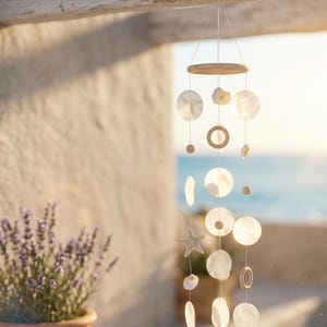 Puede incluir: Un carillón de viento de temática costera con una parte superior de madera y una serie de discos redondos de concha blanca colgantes y detalles de conchas marinas. También se incluye una concha en forma de estrella de mar. El carillón cuelga frente a una pared soleada y el mar.