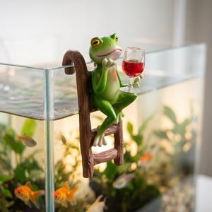 Puede incluir: Una caprichosa figura de rana verde con ojos amarillos y boca roja, sosteniendo una copa de vino tinto. La rana está posada en una escalera marrón, colocada en el borde de un acuario de vidrio lleno de peces y plantas.